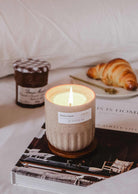 Lit reusable soy coffee candle in ceramic cup beside croissant and jar of jam on bed with book underneath