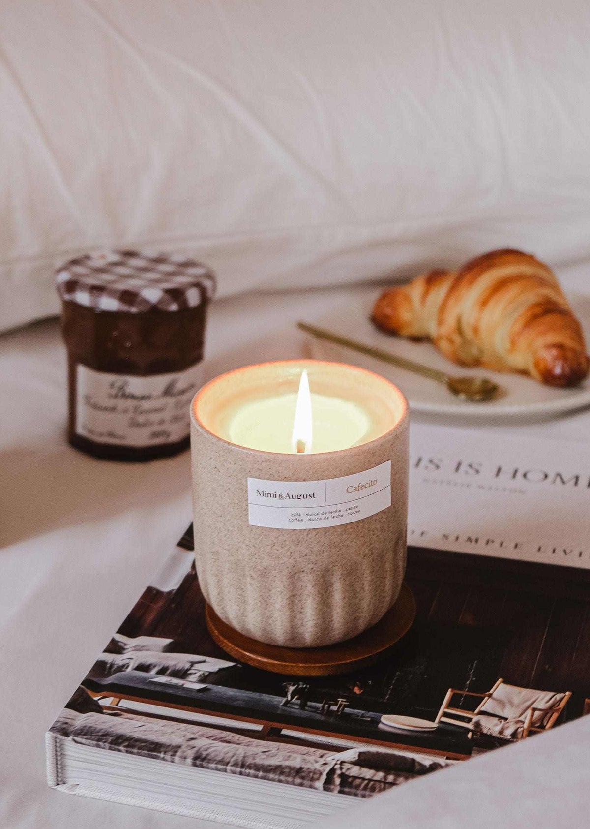 Lit reusable soy coffee candle in ceramic cup beside croissant and jar of jam on bed with book underneath