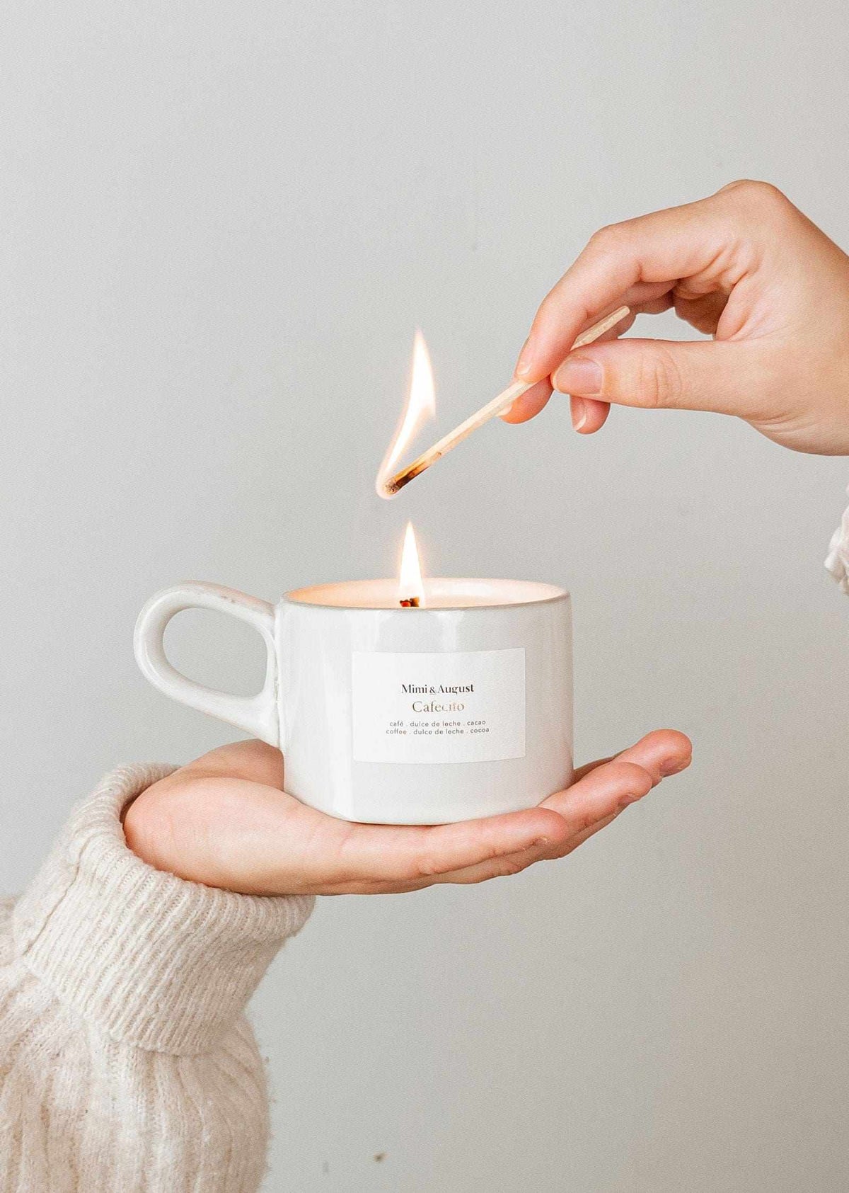 Hand lighting a reusable ceramic coffee candle in a white mug labeled Cafecito with warm flame and cozy background