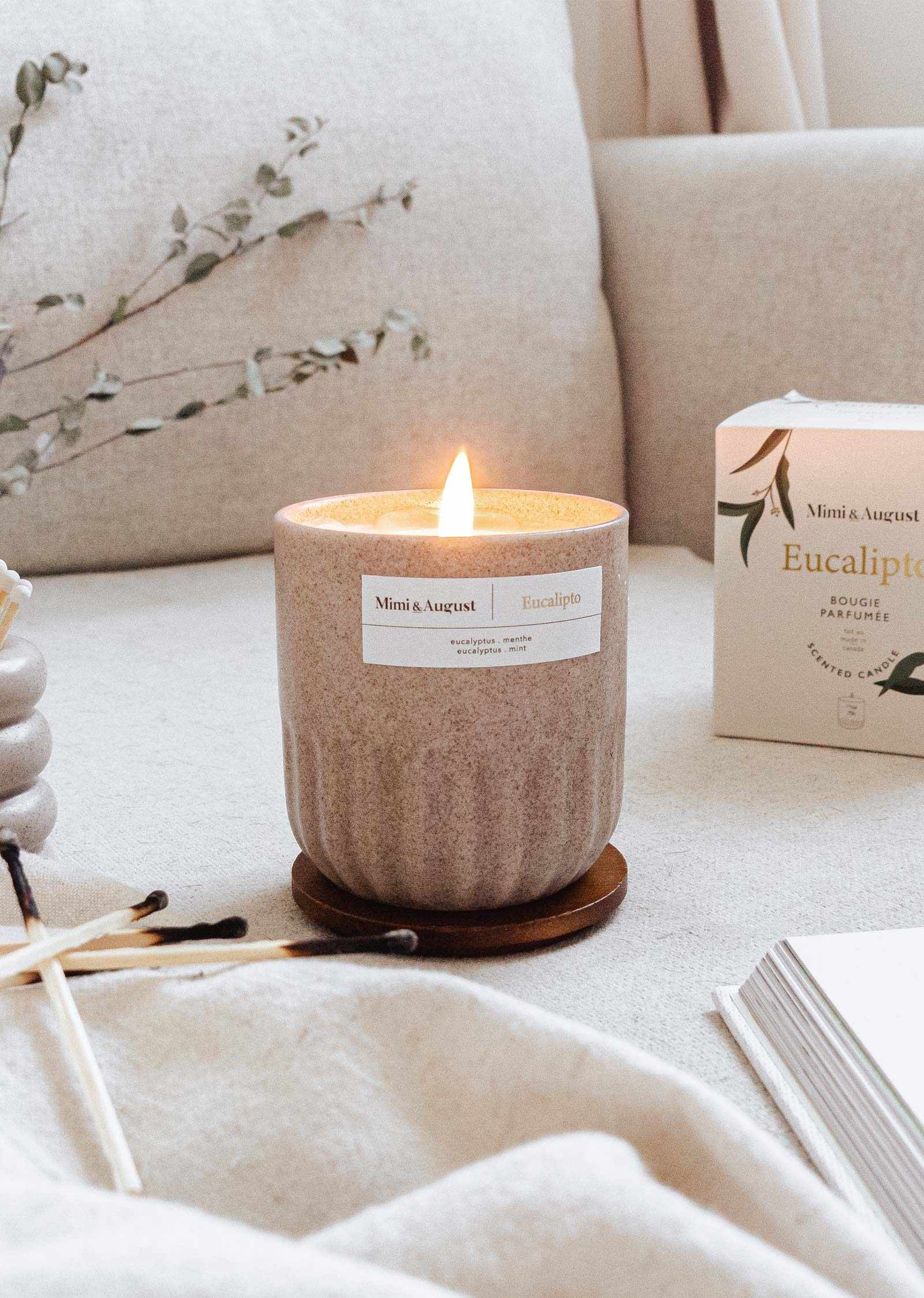 Bougie eucalyptus allumée dans une tasse en céramique réutilisable avec des feuilles d'eucalyptus en arrière-plan.