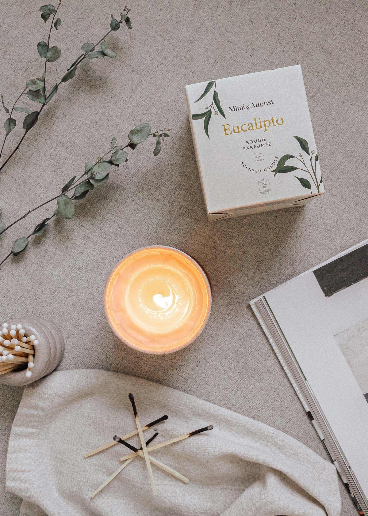 Lit eucalyptus candle in reusable ceramic holder with packaging and natural decor on beige fabric background