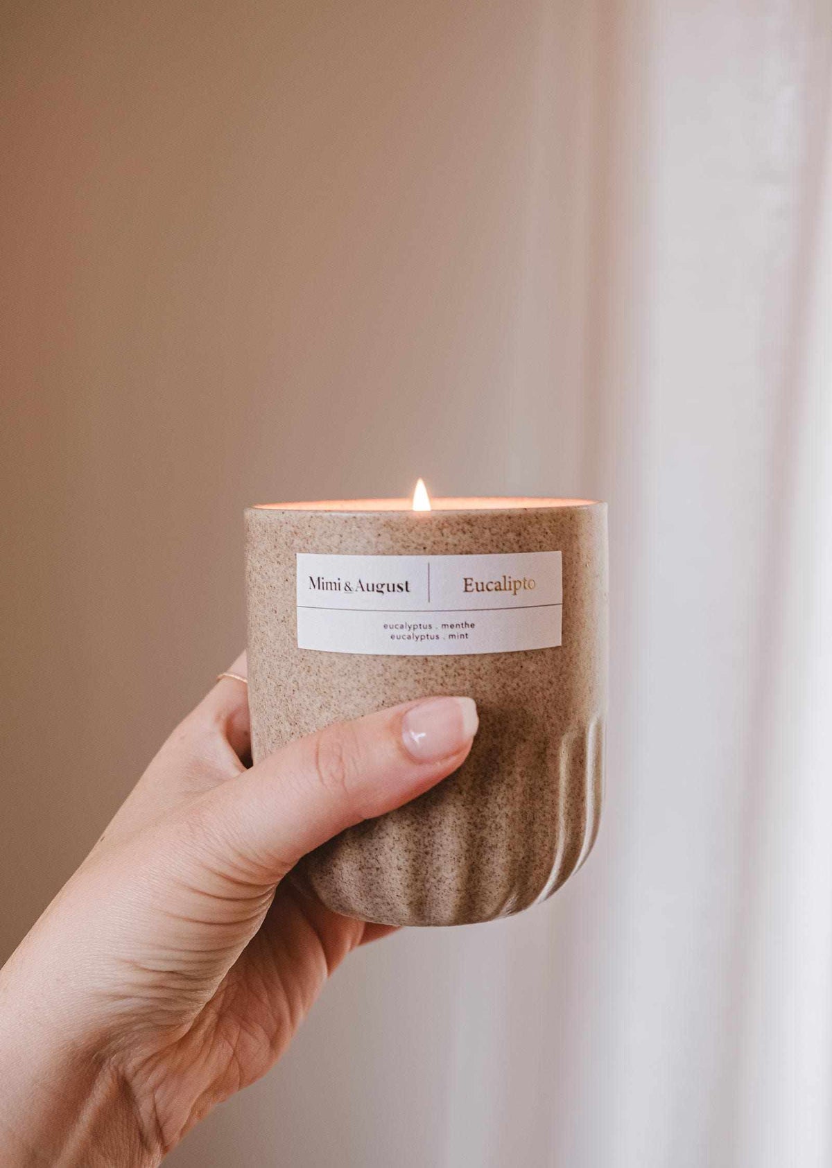 Hand holding lit eucalyptus candle in reusable ceramic cup with soft neutral background
