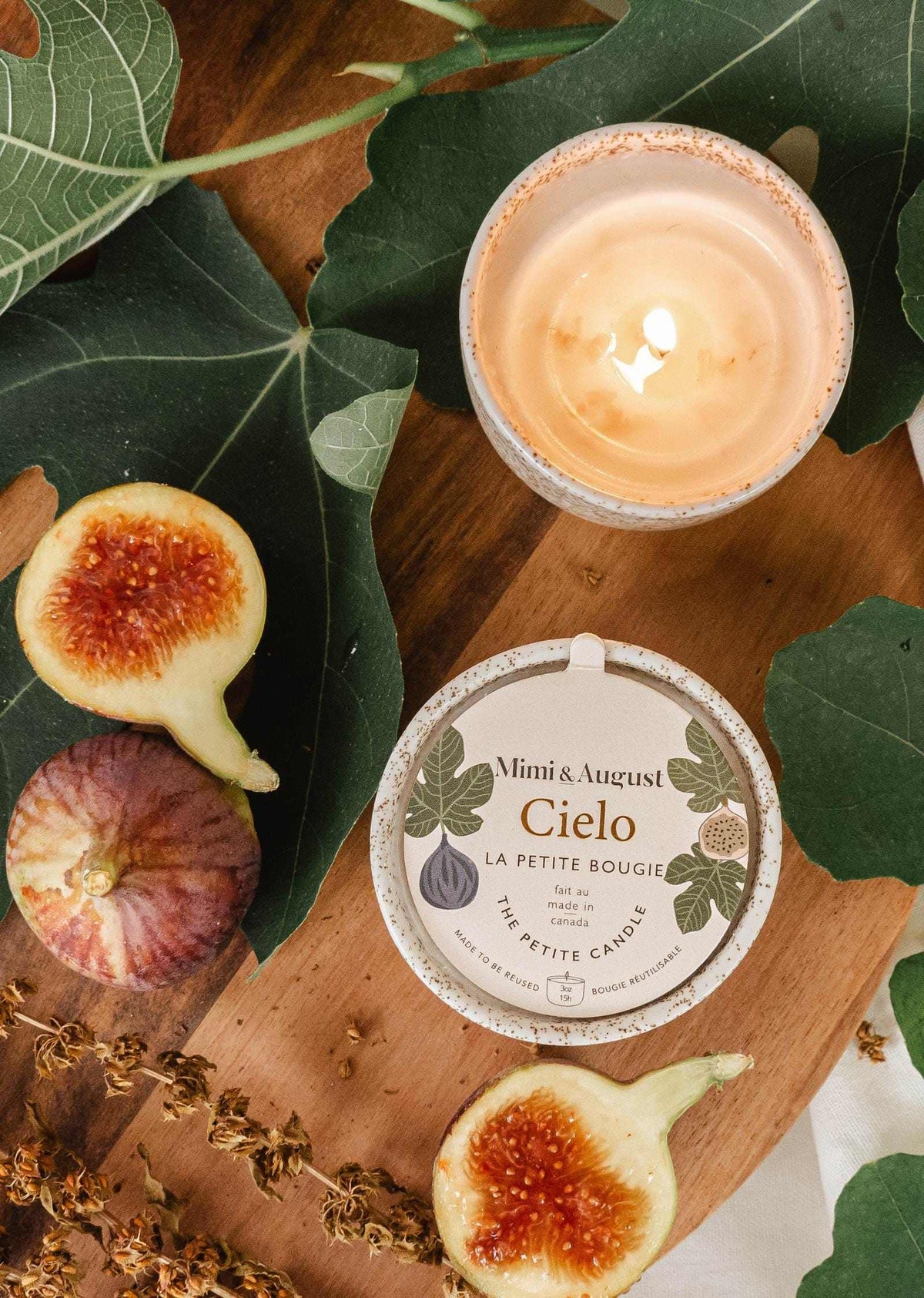Burning fig candle in reusable ceramic cup with fresh figs and green leaves on wooden surface