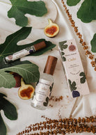A bottle and box of Mimi & August Cielo fig room spray, made with premium fragrance oil, are displayed on a white cloth decorated with fig leaves, dried flowers, and fresh figs.