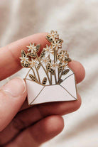 Hand holding a 38mm gold tone brass enamel pin shaped like an envelope filled with flowers, titled Letter full of flowers.