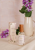 A bottle of Mimi & August's Room Spray - Lilas and its box sit on a beige surface, with a ceramic pitcher and purple flowers in the background.