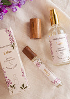 Room spray - Lilas by Mimi & August and packaging on a fabric surface with purple flowers. Includes a boxed spray, an uncapped spray bottle, a woody cap, and a small spray vial. Labeled "Mimi & August Lilas," this Room spray - Lilas features premium fragrance oil for an exquisite scent experience.