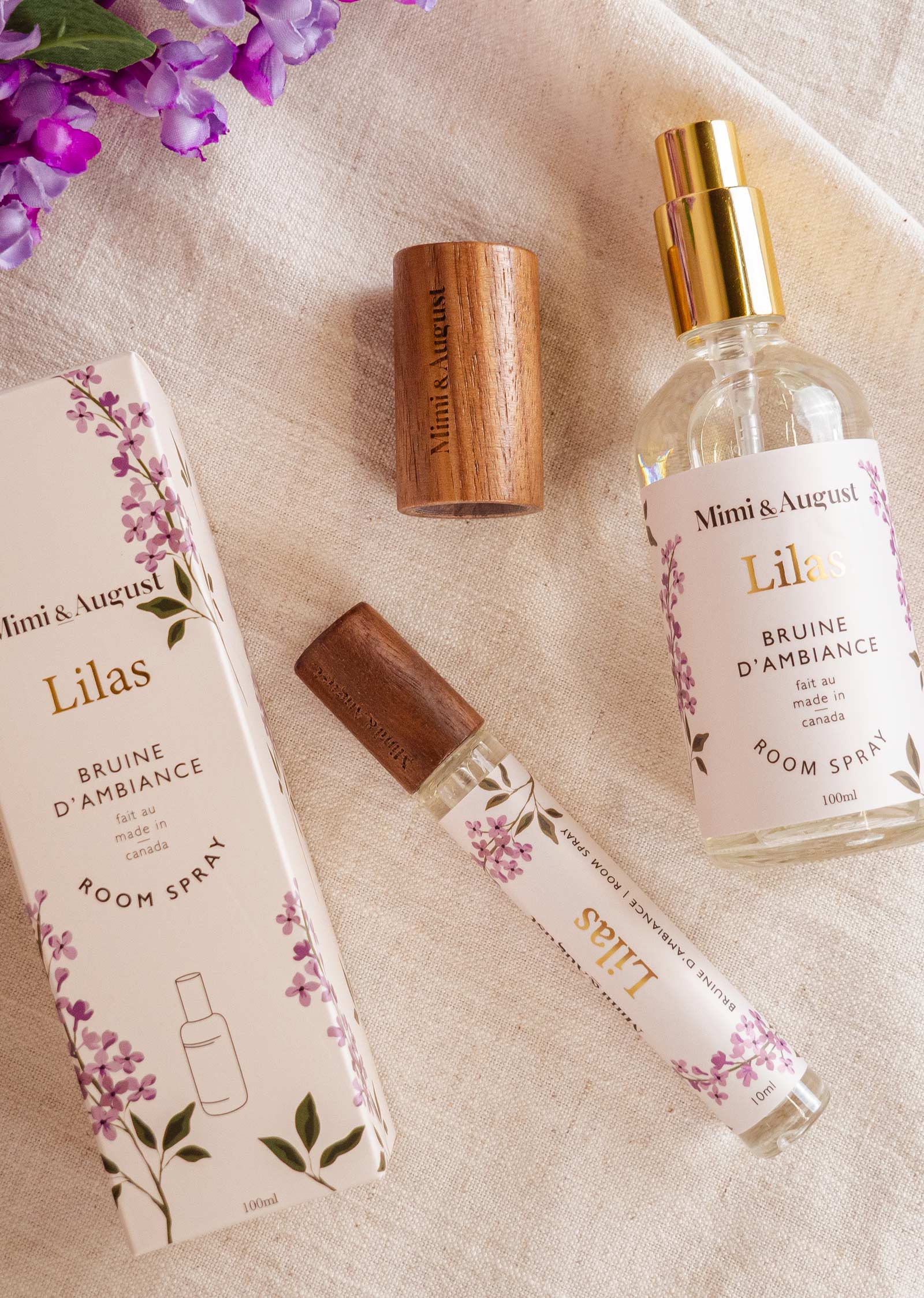 Vaporisateur d'ambiance - Lilas de Mimi & August et emballage sur une surface en tissu avec des fleurs violettes. Comprend un vaporisateur en boîte, un flacon pulvérisateur non bouché, un bouchon en bois et un petit flacon pulvérisateur. Étiqueté "Mimi & August Lilas", ce vaporisateur d'ambiance - Lilas contient une huile de parfum de première qualité pour une expérience olfactive exquise.