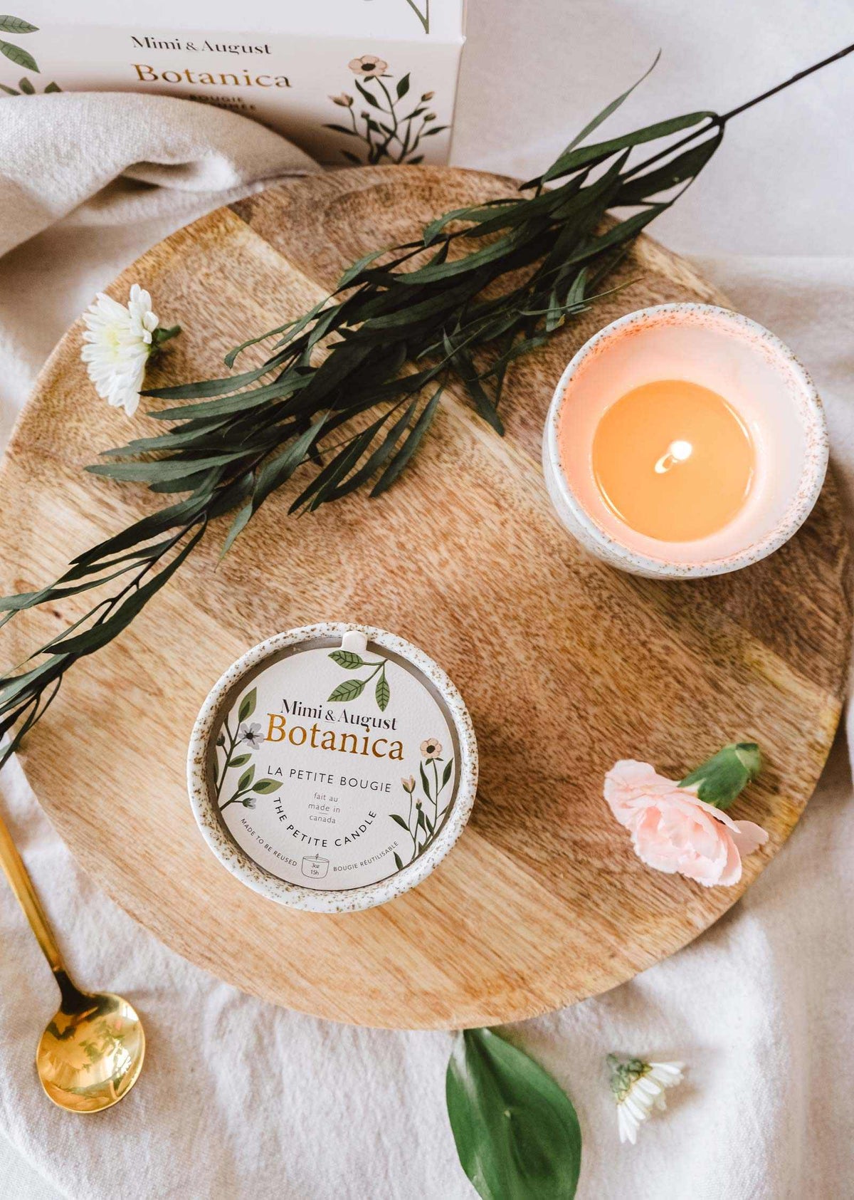 Vue à plat d'un plateau en bois avec une bougie de soya réutilisable Botanica de Mimi & August allumée, des produits de beauté et des fleurs éparpillées, évoquant une atmosphère paisible de spa.