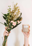 Des mains tenant un bouquet de fleurs roses et une bougie de soya réutilisable Botanica de Mimi & August sur un fond neutre.