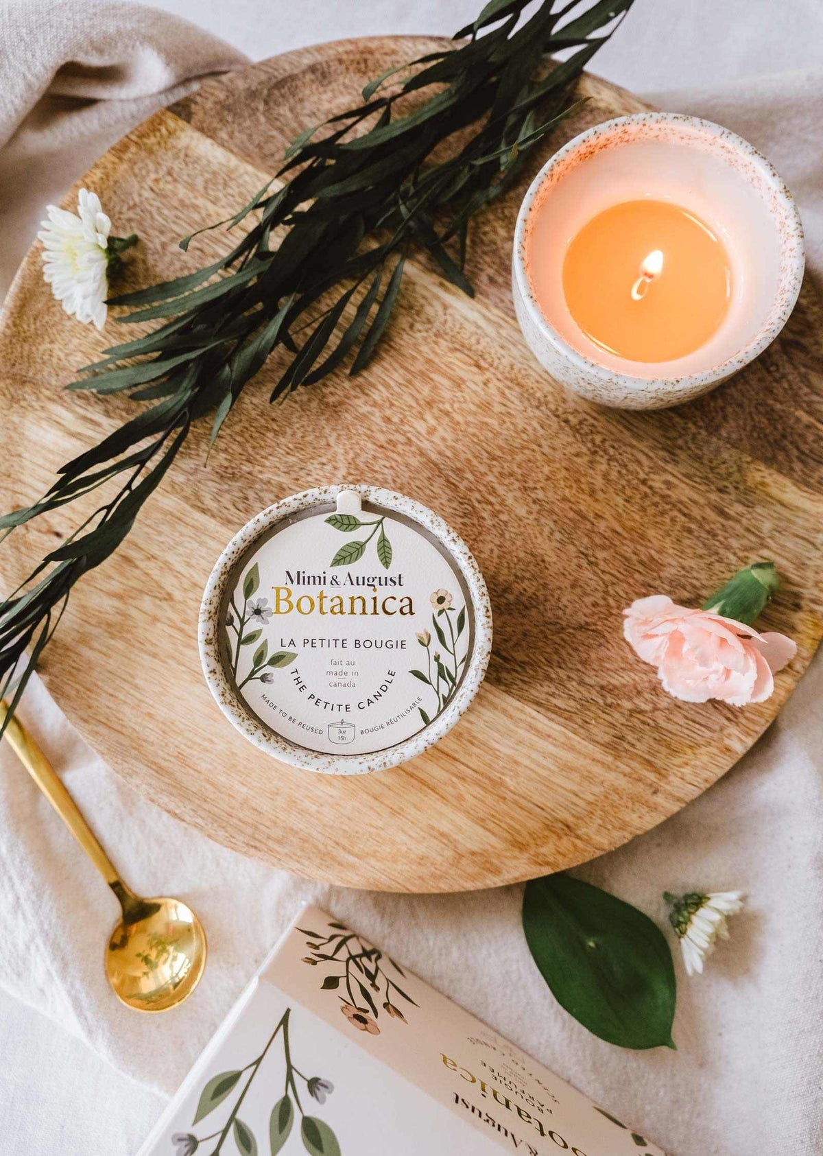 Une bougie de soya réutilisable Botanica de Mimi & August et un petit livre posés sur un plateau en bois décoré de fleurs et de feuilles vertes, créant une ambiance sereine.