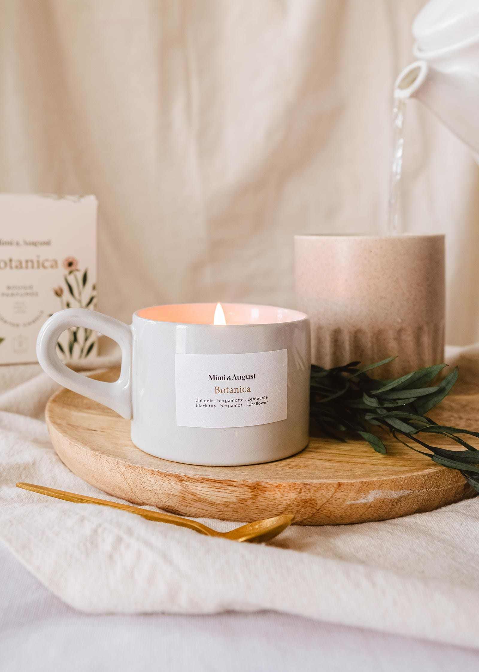 Une bougie de soya réutilisable Botanica de Mimi & August allumée sur un plateau en bois avec un brin de verdure et un objet décoratif dans un décor douillet et tamisé.