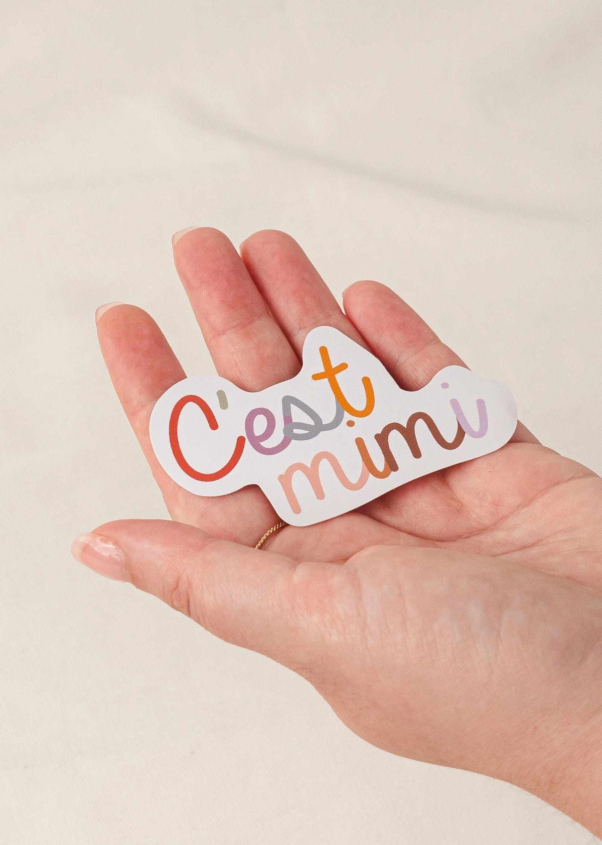 Hand holding a water-resistant vinyl sticker with "C'est mimi" text and white border