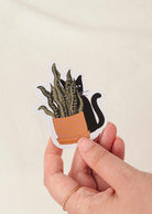Hand holding durable vinyl sticker of black cat peeking from behind a potted plant with white border